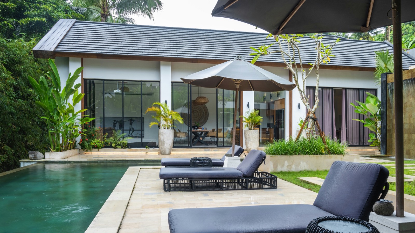 Terrasse de piscine meublée de transats et de parasols sombres, devant une villa aux larges baies vitrées